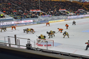 Gruppe von Menschen, die Hockey auf einer Indoor-Eisbahn spielen, umgeben von einem Zaun, unter Stadionbeleuchtung, mit Zuschauern im Hintergrund und sichtbaren Bannern.