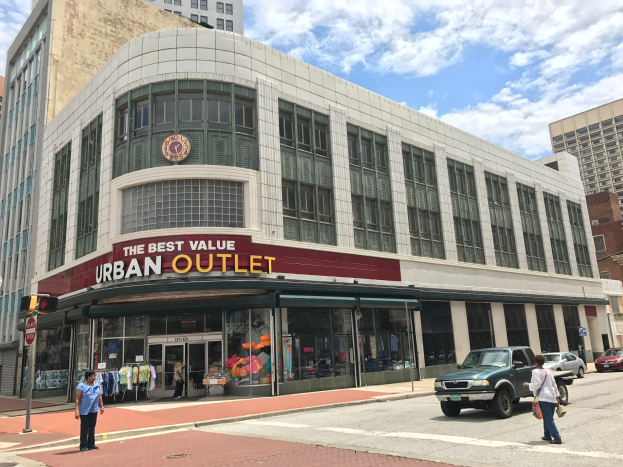 Eine belebte Stadtstraße mit Fahrzeugen auf der Straße und Fußgängern auf dem Gehweg, mit hohen Glasgebäuden und einem Schild im Hintergrund, das "The Best Value Urban Outlet" anzeigt.