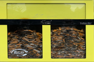 Gelber Recycling-Eimer voller Zigarettenstummel auf einem Gehweg, beschriftet mit Text.