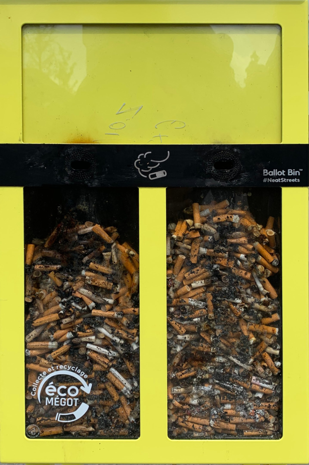 Gelber Recycling-Eimer voller Zigarettenstummel auf einem Gehweg, beschriftet mit Text.