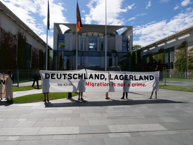 Eine Gruppe von Menschen hält ein Transparent mit der Aufschrift "Deutschland, Lagerland Migration ist kein Verbrechen" vor einem Gebäude mit Fenstern, umgeben von Gras, einem Metallzaun, Pflanzen, Bäumen und Fahnenstangen, unter einem bewölkten Himmel.