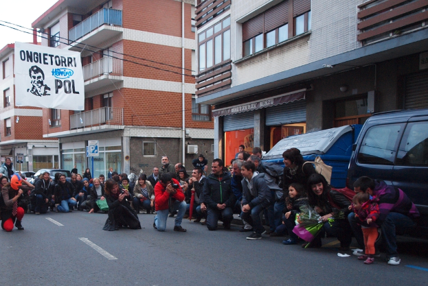 Eine Menschenmenge auf einer Stra├če, mit zwei Individuen, die Kameras an der Front halten, einem Auto auf der rechten Seite, Geb├Ąuden mit Fenstern im Hintergrund und einem Banner auf der linken Seite.