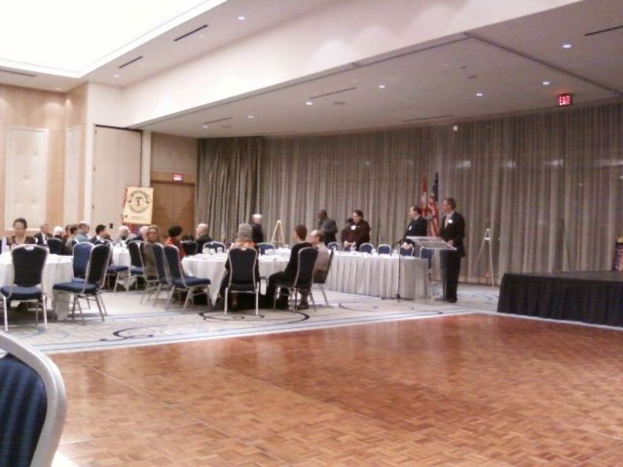 Eine Gruppe von Menschen, die auf Stühlen sitzen und einige stehen, umgeben von Fahnen auf Stangen, Lichtern, Tafeln und Vorhängen im Hintergrund.