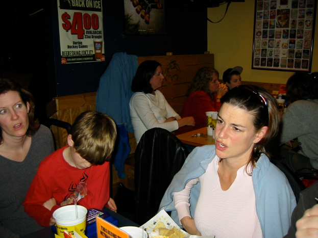 Frauen an einem Tisch sitzend mit Flaschen, Gläsern und Lebensmitteln, mit Plakaten und einem gerahmten Bild an der Wand dahinter.