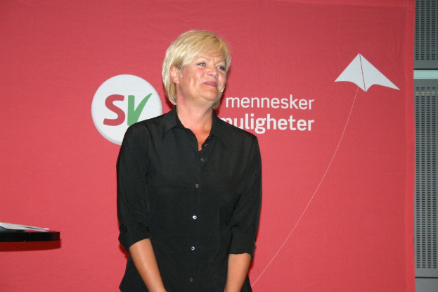 Eine Frau mit einem Hemd und einem Mikrofon steht vor einer Tafel, mit Papieren auf einem Tisch links von ihr und einem Stahlfenster rechts von ihr.