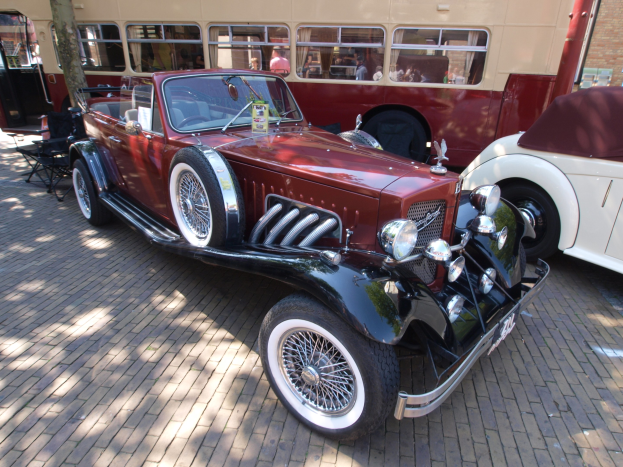 Ein Oldtimer-Auto neben einem Doppeldeckerbus auf einem Kopfsteinpflasterweg geparkt, mit Stühlen und einem Baum im Vordergrund, einem Gebäude mit Fenstern im Hintergrund und Menschen im Bus zu sehen.