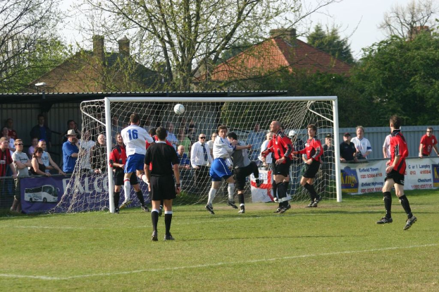 Spieler spielen Fussball auf einem Feld mit einem Tor, hinter dem Zuschauer stehen, mit B├Ąumen und H├Ąusern im Hintergrund.