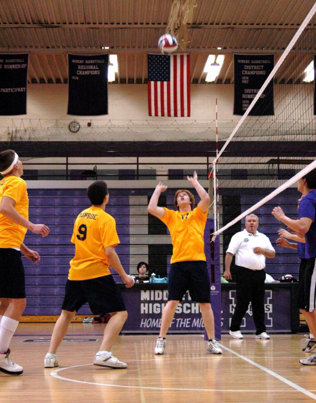 Menschen stehen auf einem Spielfeld mit Netz und Ball, tragen Kleidung und Schuhe, mit einem Landesflagge, Wand und Postern mit Text im Hintergrund.