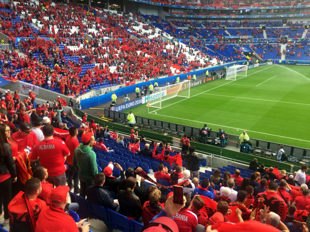 Ein volles Stadion mit sitzenden und stehenden Zuschauern, Bannern, einem Torpfosten und Deckenleuchten, während die Menge gespannt auf den Beginn des Fußballspiels wartet.
