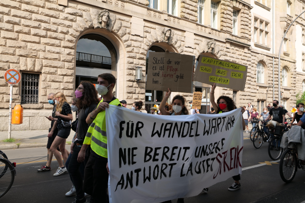 Eine Gruppe von Menschen marschiert auf der Straße in Berlin, mit Schildern und Transparenten, einige fahren Fahrräder, vor einem Gebäude mit Bögen, Säulen, Skulpturen und Bäumen.