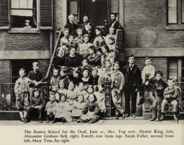 Eine Gruppe von Menschen, die vor einem Gebäude mit Fenstern und einer Tür stehen und sitzen, mit Text unten, der "The Boston School for the Deaf, June 21, 1871" lautet.