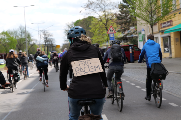 Eine Gruppe von Menschen, die auf Fahrrädern eine Straße entlangfahren, mit einer Frau vorn, die ein Schild mit der Aufschrift "Exit Racism" hält.