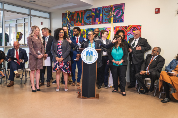 Gruppe von Menschen vor einem Rednerpult mit Mikrofon und einer Tafel mit dem Text "Sancho City Councilman Joins City Council". Im Hintergrund sind Flaggen, eine Tafel, ein Feuerlöscher und einige Deckenleuchten zu sehen.