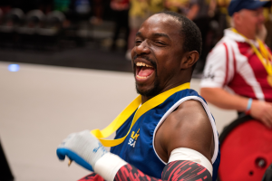 Ein Mann im Rollstuhl mit ausgestreckten Armen und einer Medaille um den Hals lächelt im Vordergrund, mit Menschen, Stühlen und anderen Gegenständen im Hintergrund.
