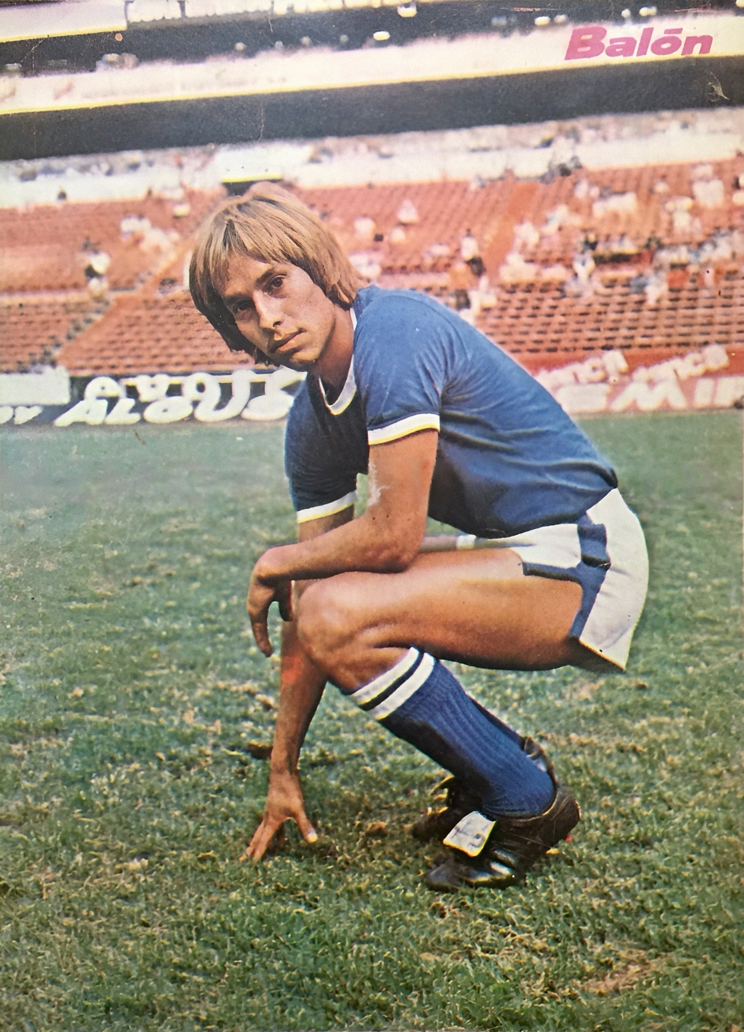 Ein Mann kniet auf einem Fußballfeld umgeben von Menschen auf Stühlen und Schildern mit Text, trägt Schuhe.