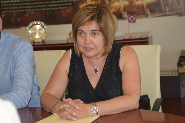 Eine Frau in einem schwarzen Oberteil sitzt auf einem Stuhl und schaut geradeaus, neben ihr ein Mann in einem blauen Hemd; ein Award-Trophy und ein gelbes Plakat sind im Hintergrund sichtbar.