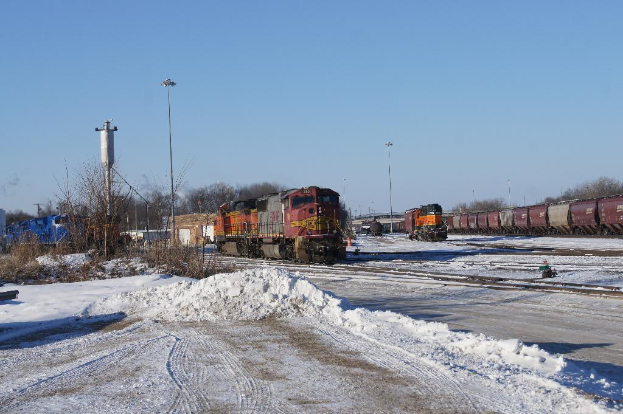 Eine Lokomotive vorne mit einem Zugwagen auf der rechten Seite auf Schienen geparkt, Schnee im Vordergrund und trockene Bäume mit einem Strommast im Hintergrund.