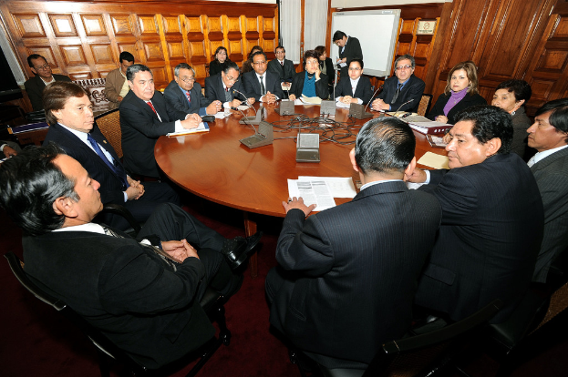 Eine Runde-Tisch-Sitzung mit Delegierten, die ├╝ber Mikrofone, Papiere, Kabel und Dateien auf dem Tisch sprechen, mit einer h├Âlzernen Wand im Hintergrund.