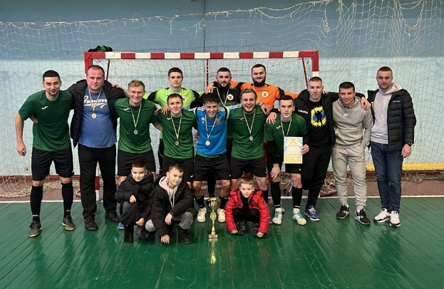 Eine Gruppe von Männern in Medaillen, einer hält einen Pokal, steht auf einem Futsal-Platz mit einem Tor und einer Wand im Hintergrund und feiert ihren Futsal-Weltmeisterschaftssieg.