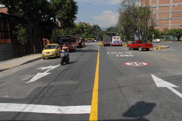 Eine Straßenszene mit Gebäuden, Bäumen, fahrenden Fahrzeugen, Straßenrandtafeln, Verkehrsampeln, Warnmarkierungen und einem blauen bewölkten Himmel.