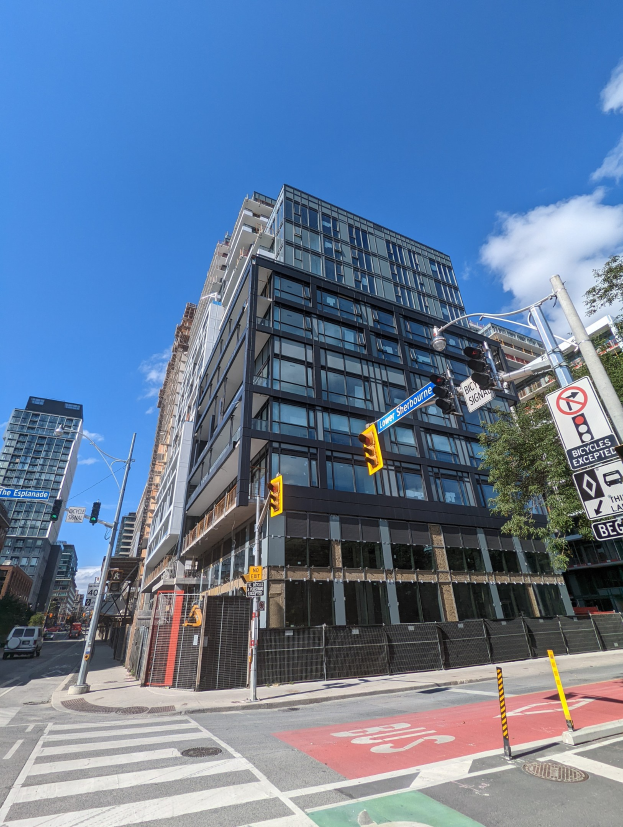 Eine belebte Stadtstraße mit Fahrzeugen, Verkehrsampeln, Schildern, Bäumen und einem Zaun, im Hintergrund ein hoher Wohnkomplex mit der Aufschrift "541 East 57th Street, Unit 541" unter einem bewölkten Himmel.