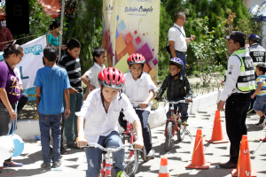Eine Gruppe von Kindern fährt Fahrräder auf einer Straße mit Verkehrskegeln, einige tragen Helme, andere stehen herum, ein Banner mit Text, Bäume und Gebäude im Hintergrund.