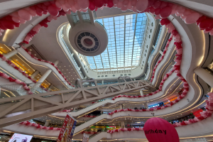 Ein Einkaufszentrum im Valentinstag-Dekor mit pinken und weißen Ballons, Rolltreppen, Geländern, Texttafeln, Lichtern und einer Glasdecke.