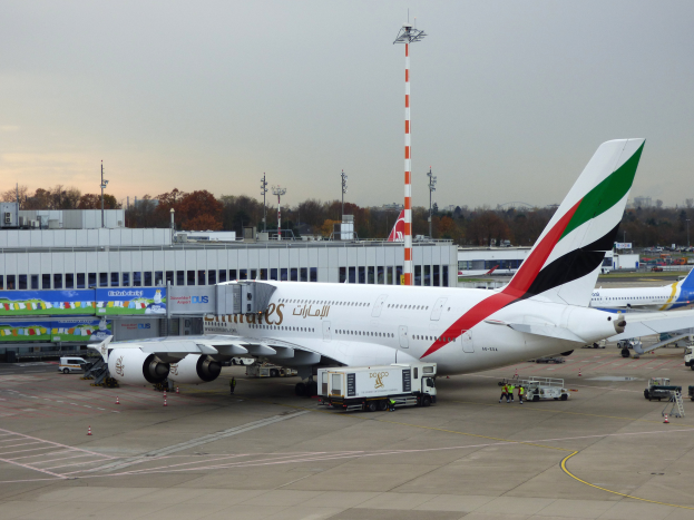 Ein Emirates Airbus A380-800 steht auf dem Frankfurter Flughafen, umgeben von Fahrzeugen und Menschen, mit Gebäuden, Bäumen, Pfählen und einem klaren blauen Himmel im Hintergrund.