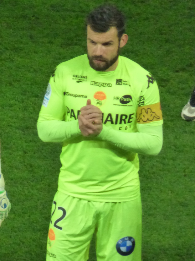 Ein Torwart in einem gelben Trikot klatscht auf einem Fußballfeld, trägt ein grünes Hemd und Shorts und hat ein Teil eines anderen Beins im Bild.