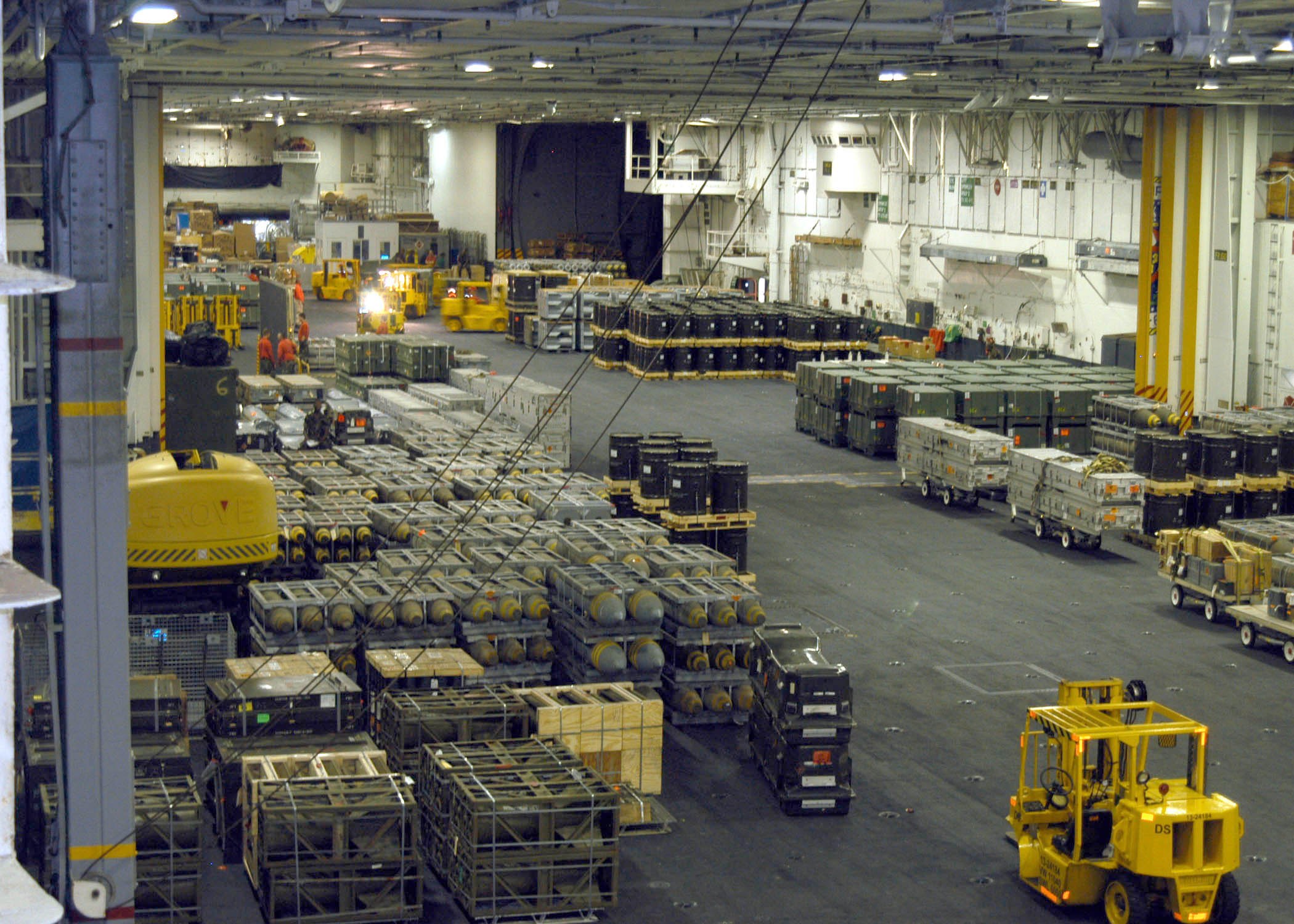 Großes Lagerinnere mit Lastwagen, Containern, Menschen, Leuchten, Metallstangen und einem sichtbaren Rad.