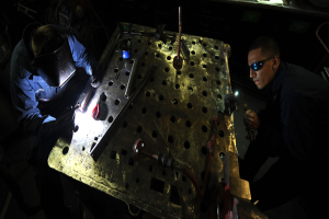 Zwei Männer in Schutzausrüstung arbeiten an einem Metallstück in einer dunklen Werkstatt, umgeben von verschiedenen Werkzeugen und Materialien auf einem Tisch.