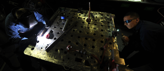 Zwei Männer in Schutzausrüstung arbeiten an einem Metallstück in einer dunklen Werkstatt, umgeben von verschiedenen Werkzeugen und Materialien auf einem Tisch.