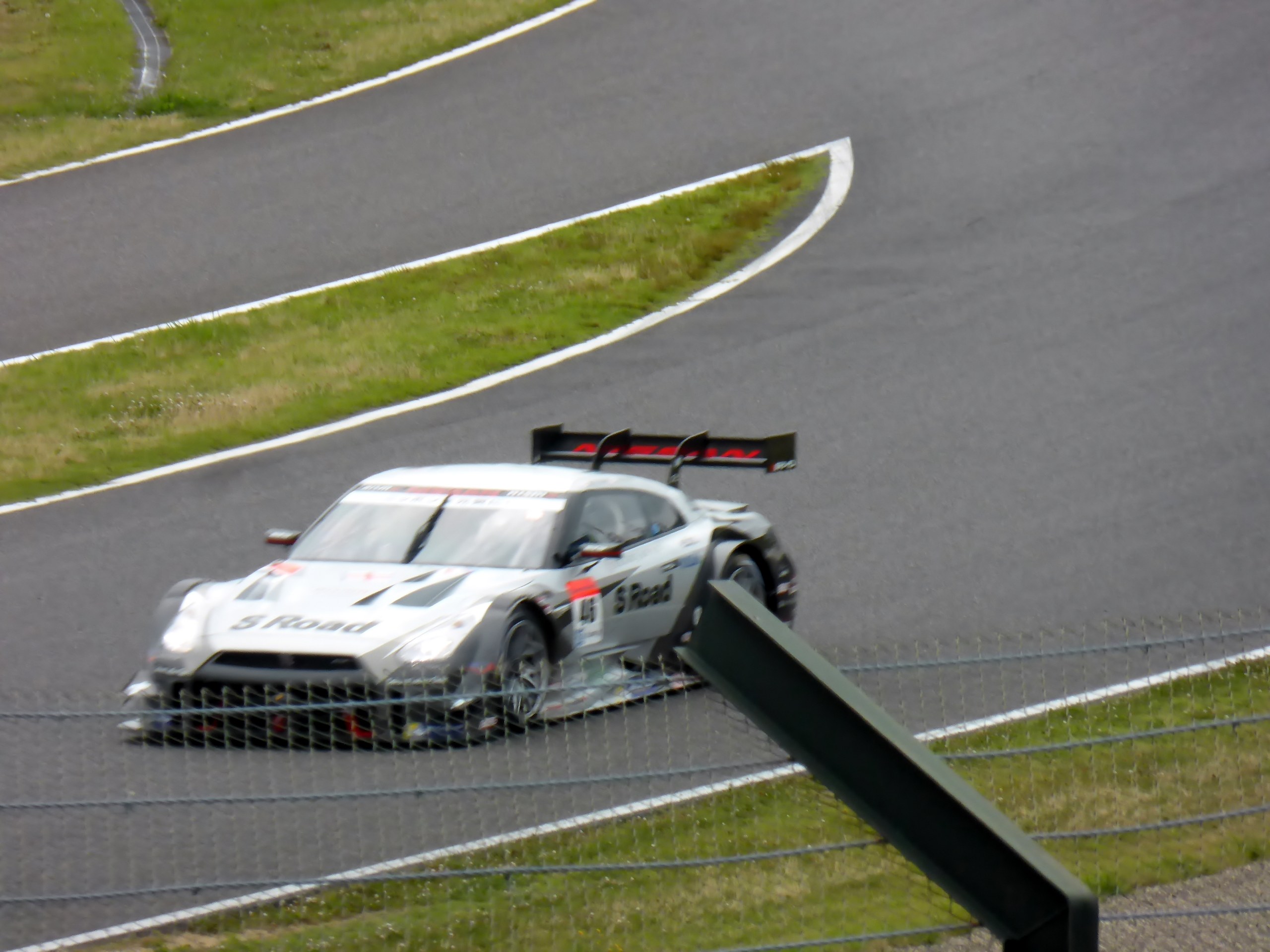 Audi R8 LMS GT3 auf der Nürburgring 24 Stunden Rennstrecke, umgeben von einem Metallzaun und saftigem Grün, in der Mitte positioniert mit der Strecke, die sich vor ihr erstreckt.