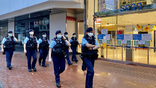 Eine Gruppe von Polizisten in Schutzmasken geht eine Straße vor einem Glasgebäude entlang, rechts davon ein Geschäft mit Plakaten an den Glaswänden.