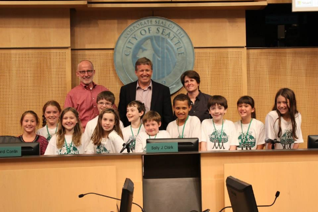 Gruppe von Kindern und Erwachsenen, die für ein Foto posen, bei einer Stadtratssitzung mit Podium, Monitoren, Mikrofonen, Logo an der Wand und einem Bildschirm im Hintergrund.