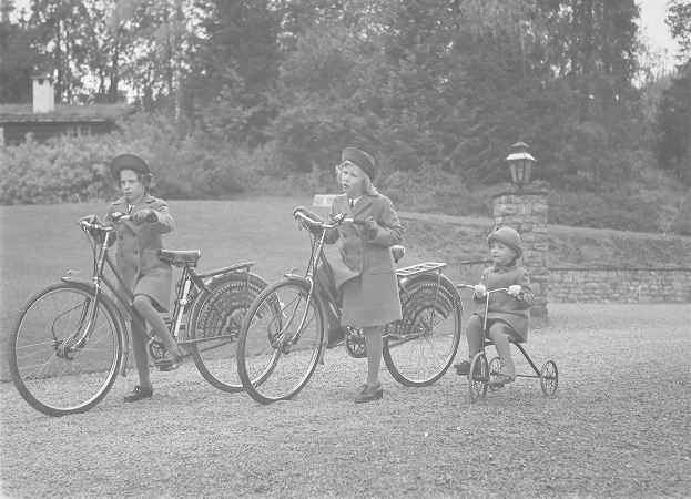 Drei Kinder fahren Fahrräder in einem Park, tragen Mäntel und Mützen, mit einem auf einem Dreirad, vor einem Hintergrund aus Bäumen, Pflanzen, einem Haus und einem Laternenpfahl, in Schwarz-Weiß.