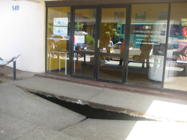 Eine gebrochene Oberfl√§che mit einer Glast√ľr, Wand und Pfahl im Hintergrund, die sich bewegende Fahrzeuge und einige stehende Personen spiegeln.