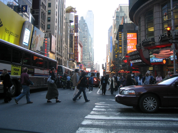 Eine belebte Stadtstraße mit Fahrzeugen, Fußgängern an einer Zebrastreifen, Gebäuden, Schildern, Verkehrszeichen und einem klaren blauen Himmel.