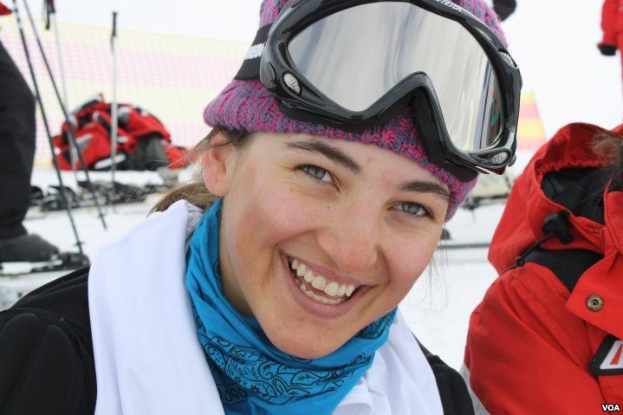 Eine Frau in einer pinken Mütze, einer Goggle, einem schwarzen und weißen Kleid und einem blauen Schal lächelt in die Kamera und steht neben einer Person in einer roten Jacke, mit Ski-Ausrüstung und einem Gebäude im Schnee im Hintergrund.