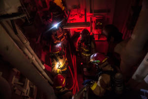 Eine Gruppe von Feuerwehrmännern in Helmen und Sauerstoffflaschen steht in einem dunklen Raum mit Rohren auf der linken und rechten Seite, mit einer Maschine und anderen Gegenständen im Hintergrund, was auf einen Rettungseinsatz hinweist.