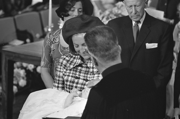 Ein Schwarz-Weiß-Foto einer Frau, die ein Baby hält, umgeben von einer Gruppe von Menschen bei einer Beerdigung, mit Stühlen und einem Tisch im Hintergrund.