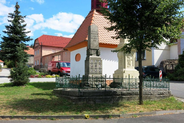 Ein kleiner Stadtplatz mit einem Holocaust-Mahnmal in der Mitte, umgeben von einem Metallzaun, umgeben von Gras, Pflanzen, Bäumen, Fahrzeugen, einem Schild, Häusern und einem bewölkten Himmel.