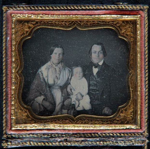 Ein Mann im Anzug und Krawatte, eine Frau mit einem Baby, alle mit ernsten Gesichtern bis auf das zufriedene Baby, in einem alten Daguerreotyp in einem Foto Rahmen.