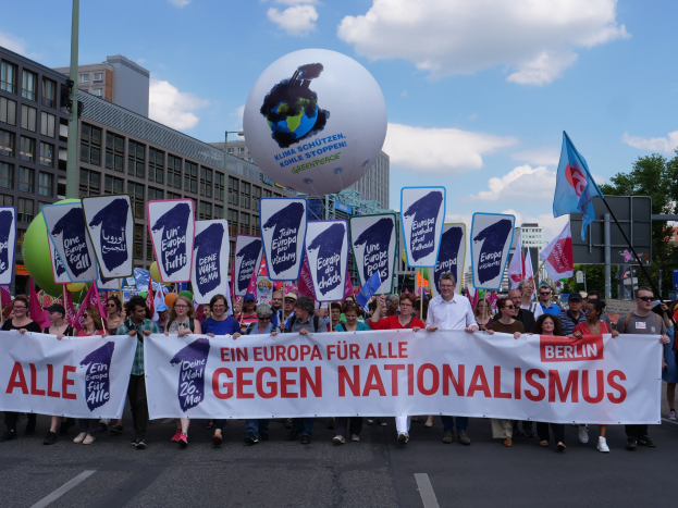 Eine Gruppe von Menschen, die mit einem Banner 'Ein Europa für alle gegen Nationalismus' und Fahnen auf Stangen die Straße entlanggehen, mit Gebäuden, Bäumen und einem bewölkten Himmel im Hintergrund.