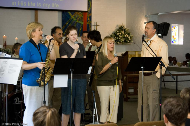 Eine Gruppe von Menschen versammelt sich in einem Raum mit einer Wandbanner, Kerzen, Papier, Mikrofonen, einem Foto Rahmen und Blumen.