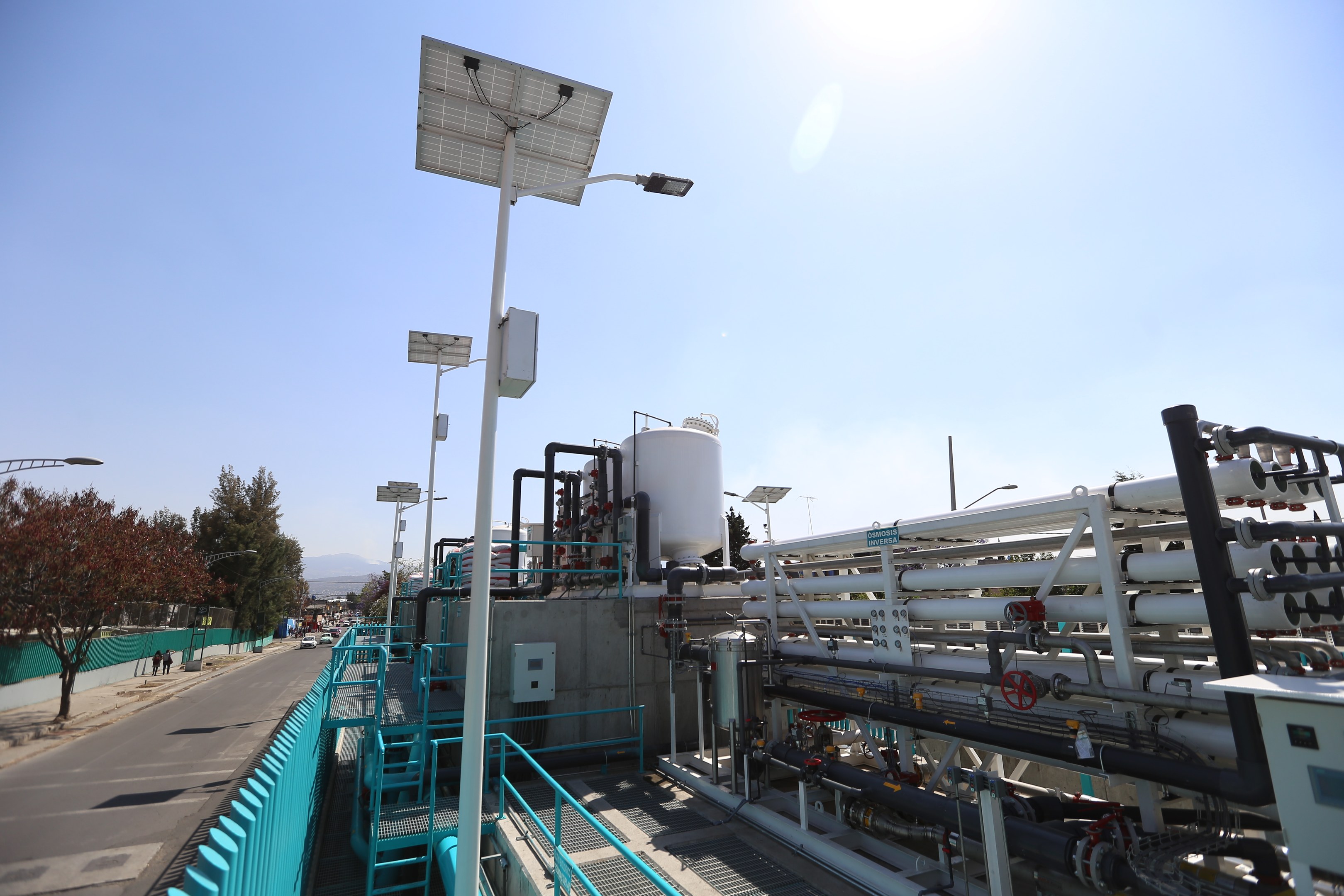 Großes industrielles Gebäude mit umfangreichen Rohrleitungen und Solarpaneelen auf dem Dach, umgeben von Sicherheitsgeländern und Infrastruktur, vor einem klaren blauen Himmel und Hügeln in Kalifornien