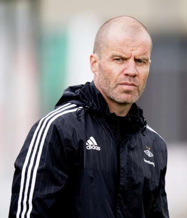 Ein kahlgeschorener Mann in einer schwarzen Jacke steht auf einem Fussballfeld und tr├Ągt eine marineblaue Jacke mit wei├čen Streifen und einem wei├čen Kragen, die H├Ąnde sind verschr├Ąnkt und er hat einen ernsten Gesichtsausdruck.