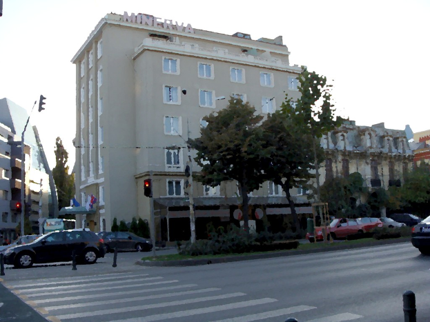 Eine befahrene Straße mit fahrenden Fahrzeugen, eine grüne Mittelinsel, Verkehrsampelmasten, Straßenlaternen, Bäume, Gebäude im Hintergrund und einen klaren Himmel darüber.
