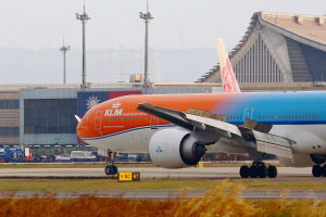 Ein KLM Boeing 777-300ER auf der Rollbahn am Frankfurter Flughafen mit Gras und Pflanzen im Vordergrund, Gebäuden und Fahrzeugen im Hintergrund und einem klaren blauen Himmel.