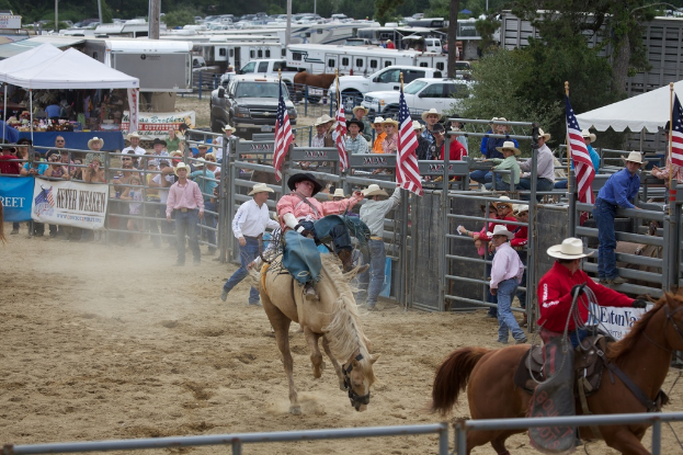 Zwei Reiter bei einem Wettbewerb namens Charreada, umgeben von Menschen, Banner, Fahnen, Zelten, geparkten Fahrzeugen und Bäumen im Hintergrund.